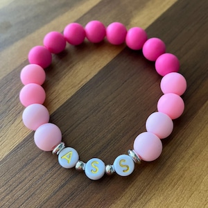 May include: A pink beaded bracelet with the letters "ASS" spelled out in white beads with gold lettering. The bracelet features a gradient of pink beads, transitioning from light to dark pink. The bracelet is on a brown wooden surface.