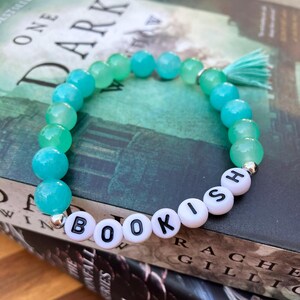 Puede incluir: Una pulsera de cuentas turquesas y verdes, con la palabra "BOOKISH" escrita con cuentas de letras blancas. Una pequeña borla turquesa está adjunta. La pulsera está sobre un libro.