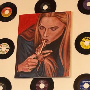 Op de afbeelding: Een olieverfschilderij van een vrouw met lang rood haar die een sigaret aansteekt. Het schilderij staat op een canvas en is omringd door vintage vinylplaten. De vrouw draagt een donkerblauwe top en een zwarte coltrui.