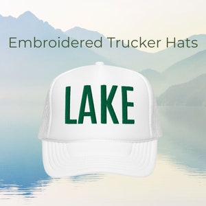 Może przedstawiać: Biała czapka trucker z haftowanym zielonym napisem "LAKE" z przodu.