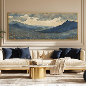 Puede incluir: Una gran pintura rectangular de acuarela de un paisaje montañoso en tonos azules y verdes, enmarcada en oro. La obra de arte cuelga sobre un sofá color crema con cojines azul marino. Una mesa de centro dorada y una manta beige completan la escena.