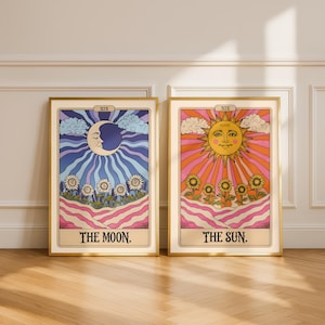 Puede incluir: Dos impresiones enmarcadas de cartas del tarot. Una presenta una luna creciente con una cara, rodeada de rayos azules y morados, y el texto "THE MOON". La otra representa un sol sonriente con rayos naranjas y el texto "THE SUN."