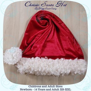 May include: A red Santa hat with a white fluffy trim. The hat is made of a soft, velvety material. Text on the hat reads "Chrissie Santa Hat By Happy Sweet Saving". The hat is available in children's and adult sizes.