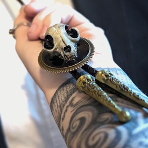 Puede incluir: Un corbatín bolo con una calavera blanca detallada con ojos negros, montada en un marco redondo de metal negro y dorado. La corbata está unida a un cordón negro con dos puntas de metal dorado.