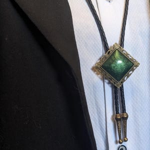 Peut inclure: Une cravate bolo avec un pendentif vert foncé en forme de losange. Le pendentif est encadré dans un métal décoratif de couleur or antique. Le cordon en cuir noir est visible sur une chemise blanche et un blazer noir.