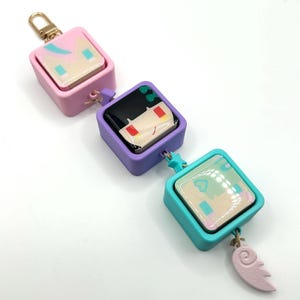May include: A keychain featuring three square charms in pink, purple, and teal, each with a unique cartoon face design. The teal charm has a pink wing-shaped dangle. The keychain has a gold-colored clasp.