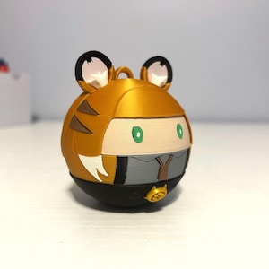 May include: A spherical figurine with a tiger-themed design. The top half is gold with black tiger stripes and ears. The bottom half is black. The figurine has green eyes and a gray and black outfit.