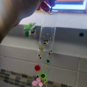 May include: A clear glass bookmark with pressed flowers and leaves. The flowers are pink, red, green, and yellow. There are also small black and yellow dots on the bookmark.