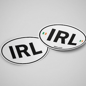 Op de afbeelding: Twee witte ovale stickers met zwarte tekst "IRL". Eén sticker heeft het woord "Ireland" eronder. Beide hebben een zwarte rand en een kleine Ierse vlag.