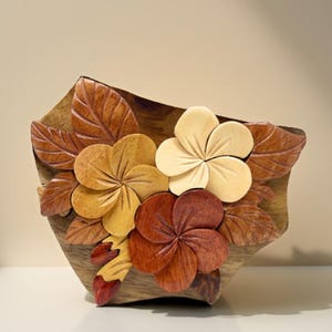 Puede incluir: Un cuenco decorativo de madera con detalles florales y de hojas talladas. Las flores son en tonos amarillo, crema y marrón rojizo, sobre un fondo de madera natural. Las hojas son de color marrón rojizo.