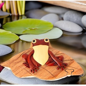 May include: A wooden puzzle box shaped like a lily pad with a red frog sitting on top. The frog has large, round eyes and a yellow belly.