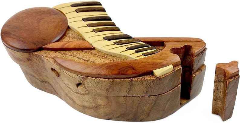 May include: A wooden piano-shaped puzzle box with a hinged lid and a separate piece that fits into the box. The box is made of light and dark wood and has a natural finish.