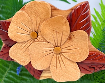 May include: A wooden puzzle box with a floral design. The box is made of light and dark wood and features two carved flowers with yellow centres. The box is surrounded by leaves.