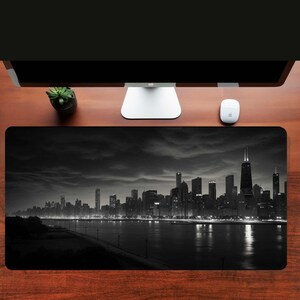 May include: Black and white photograph of a city skyline at night. The city is lit up with lights, and the water in the foreground is reflecting the lights. The sky is cloudy and dark.