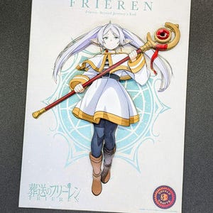 May include: A 3D printed art piece featuring the character Frieren from Frieren: Beyond Journey's End. The character has long white hair, pointed ears, and wears a white dress with gold trim, holding a staff with a red gem. Text reads Frieren.