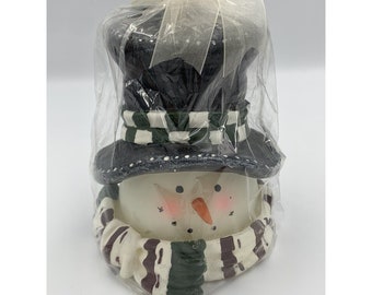 May include: A white snowman candle with a black top hat, green and white striped scarf, and a painted face. The candle is wrapped in clear plastic with a white bow.