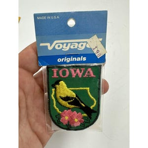 May include: A green and yellow embroidered patch with the word "IOWA" in pink letters. The patch features a yellow bird with black wings and a pink flower with yellow centers. The patch is packaged in a clear plastic bag with a blue and white label that reads "Voyage originals" and "Made in U.S.A."