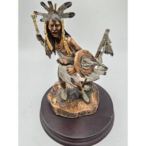 May include: A bronze sculpture of a Native American warrior holding a shield and a spear. The sculpture is on a wooden base with the words "Stand of the Sassy Warrior" inscribed on it.