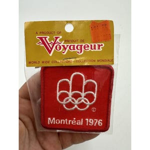 May include: Red felt patch with white embroidered Olympic rings and the text "Montréal 1976". The patch is packaged in a plastic bag with a yellow and white label that reads "A Product of Voyageur World Wide Collectors - Collection Mondiale".