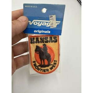 May include: A vintage Kansas Frontier West patch with a black silhouette of a rider on a horse, set against an orange and yellow background. The patch is in a clear plastic sleeve, with a blue Voyager Originals header.
