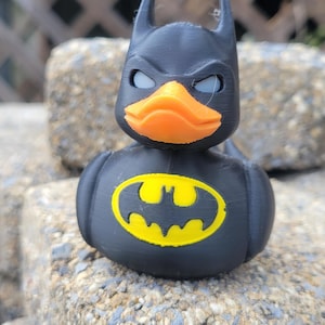 May include: A black Batman duck figurine with an orange beak and yellow Batman logo on its chest. The duck has a Batman cowl with pointed ears. The figurine is sitting on a rough, gray stone surface.