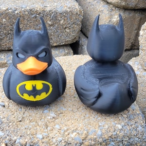 May include: Two black rubber duckies, one with a Batman logo and orange beak, the other a plain black duck. The Batman duck has a yellow logo on its chest. Both ducks have the iconic Batman cowl.