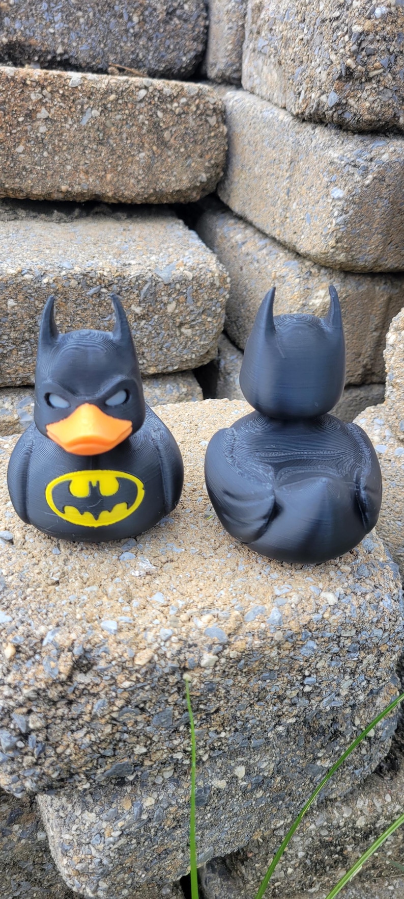 May include: Two black rubber duckies, one with a Batman logo and orange beak, the other a plain black duck. The Batman duck has a yellow logo on its chest. Both ducks have the iconic Batman cowl.