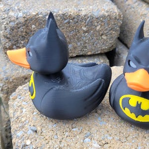 May include: Two black rubber duck toys, each with a Batman mask and logo. The ducks have orange beaks and yellow Batman symbols on their chests. They are sitting on a rough, gray stone surface.