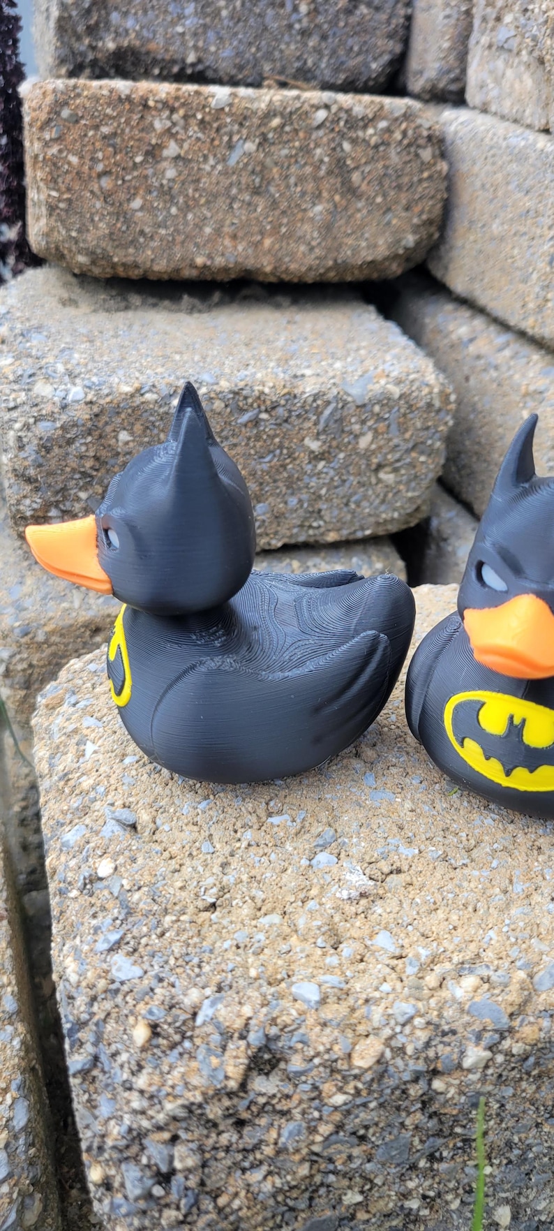 May include: Two black rubber duck toys, each with a Batman mask and logo. The ducks have orange beaks and yellow Batman symbols on their chests. They are sitting on a rough, gray stone surface.