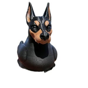 May include: A 3D-printed Doberman Pinscher figurine. The dog has a black body with tan markings on its snout, above its eyes, and on its chest. The dog's ears are erect and pointed. The figurine is sitting.