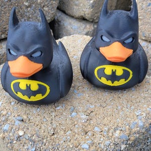May include: Two black Batman rubber ducks with orange beaks and yellow Batman logos on their chests. The ducks have pointed ears and are sitting on a rough, gray surface.