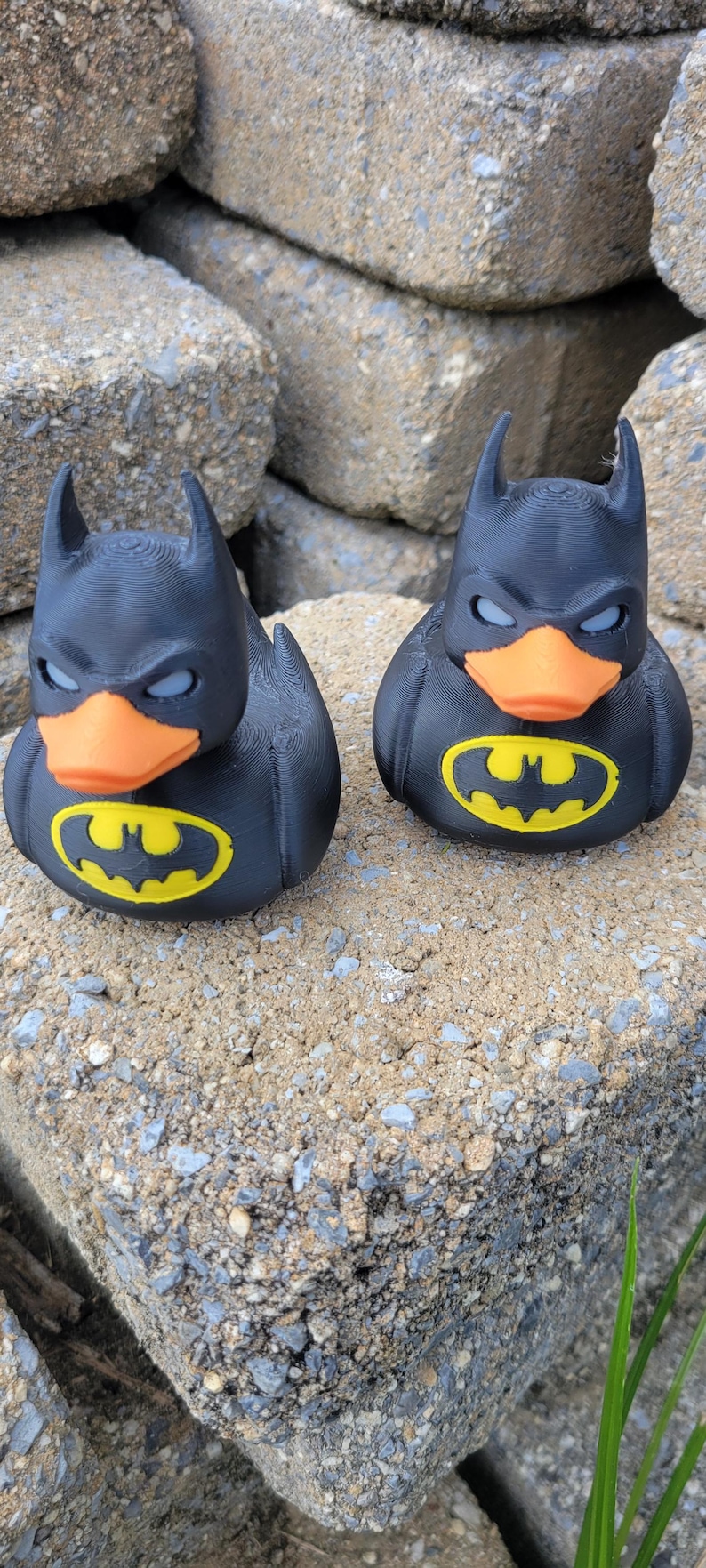 May include: Two black Batman rubber ducks with orange beaks and yellow Batman logos on their chests. The ducks have pointed ears and are sitting on a rough, gray surface.