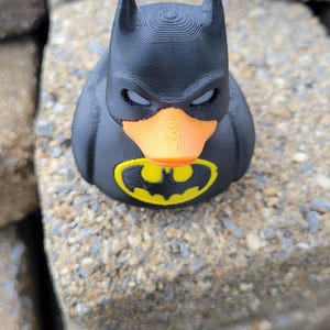 May include: A black rubber duck with a Batman mask and logo. The duck has an orange beak and the iconic yellow and black bat symbol on its chest. The toy is sitting on a stone surface.