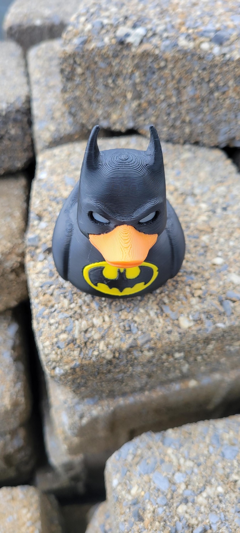 May include: A black rubber duck with a Batman mask and logo. The duck has an orange beak and the iconic yellow and black bat symbol on its chest. The toy is sitting on a stone surface.