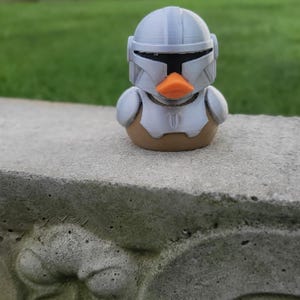 Może przedstawiać: Gumowa kaczka, zaprojektowana tak, aby przypominała postać z science fiction. Kaczka ma srebrny hełm i zbroję, z pomarańczowym dziobem. Tło jest rozmyte, z zieloną trawą i pochmurnym niebem.