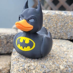 May include: A black rubber ducky with a Batman mask and logo. The duck has an orange beak, gray eyes, and a yellow and black Batman logo on its chest. The duck is sitting on a rough, gray stone surface.