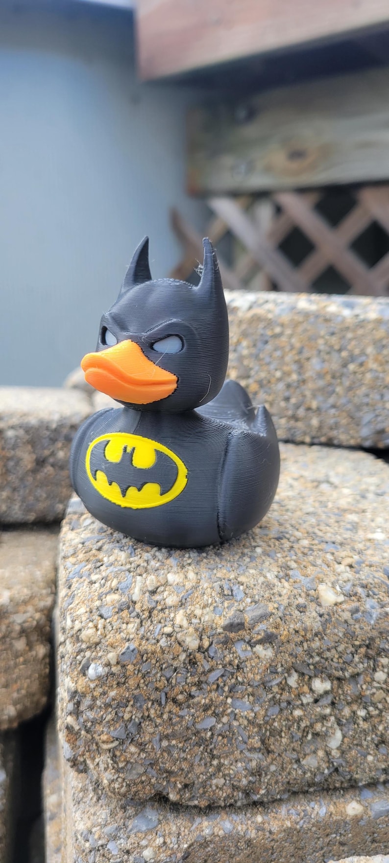 May include: A black rubber ducky with a Batman mask and logo. The duck has an orange beak, gray eyes, and a yellow and black Batman logo on its chest. The duck is sitting on a rough, gray stone surface.