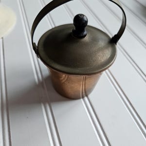 May include: A small, copper-colored bucket with a matching lid and a dark handle. The lid is topped with a small, round, black knob. The handle is arched and attached to the sides of the bucket. The bucket is sitting on a white surface.