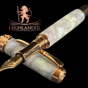 May include: A high-end fountain pen featuring a pearlescent white barrel and gold-coloured detailing. The nib is gold-coloured and has an ornate design. The pen is from Highlander Writing Instruments, as shown by the logo.
