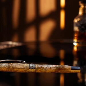 May include: A luxury fountain pen with a marbled, amber-colored barrel and silver accents. The pen is lying on a reflective surface, with a blurred bottle of amber liquid in the background. The pen's design suggests a focus on writing instruments and stationery.