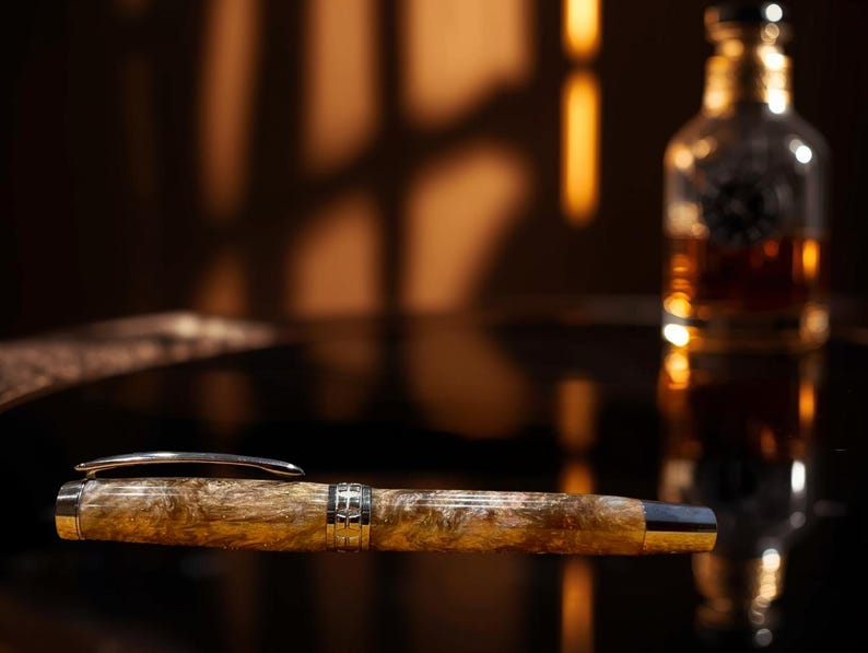 May include: A luxury fountain pen with a marbled, amber-colored barrel and silver accents. The pen is lying on a reflective surface, with a blurred bottle of amber liquid in the background. The pen's design suggests a focus on writing instruments and stationery.