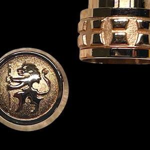 May include: Gold-coloured metal bottle stopper with a detailed design. One part features a circular emblem with a lion holding a staff. The other part has a textured, geometric pattern. The background is black.