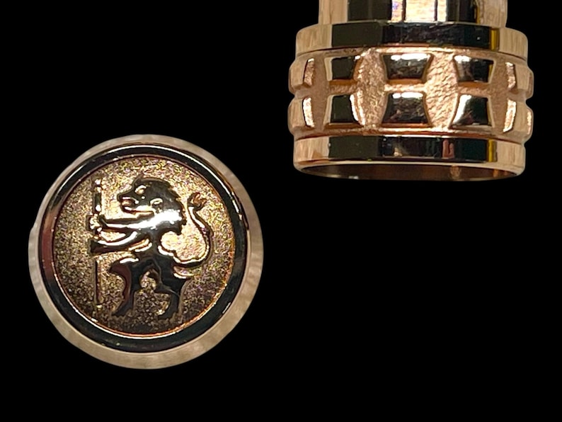 May include: Gold-coloured metal bottle stopper with a detailed design. One part features a circular emblem with a lion holding a staff. The other part has a textured, geometric pattern. The background is black.