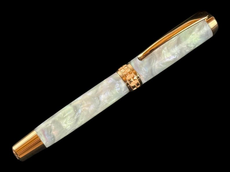 May include: A fountain pen with a pearlescent white barrel and gold-coloured accents. The pen has a gold clip and a decorative gold band near the centre. The pen's tip and end cap are also gold, creating a sophisticated writing instrument.
