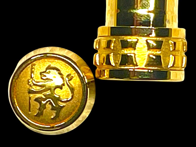Puede incluir: Primer plano de un tap&oacute;n de botella dorado con un emblema de le&oacute;n. El tap&oacute;n tiene un dise&ntilde;o detallado y texturizado. El fondo es negro, lo que resalta el color dorado del tap&oacute;n.