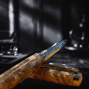 May include: A luxury pen with a silver tip and gold-toned barrel. The pen's barrel has a marbled appearance with brown and gold hues. The pen is partially uncapped, revealing the silver clip and the pen's nib. The background is blurred, with a glass and a dark bottle.