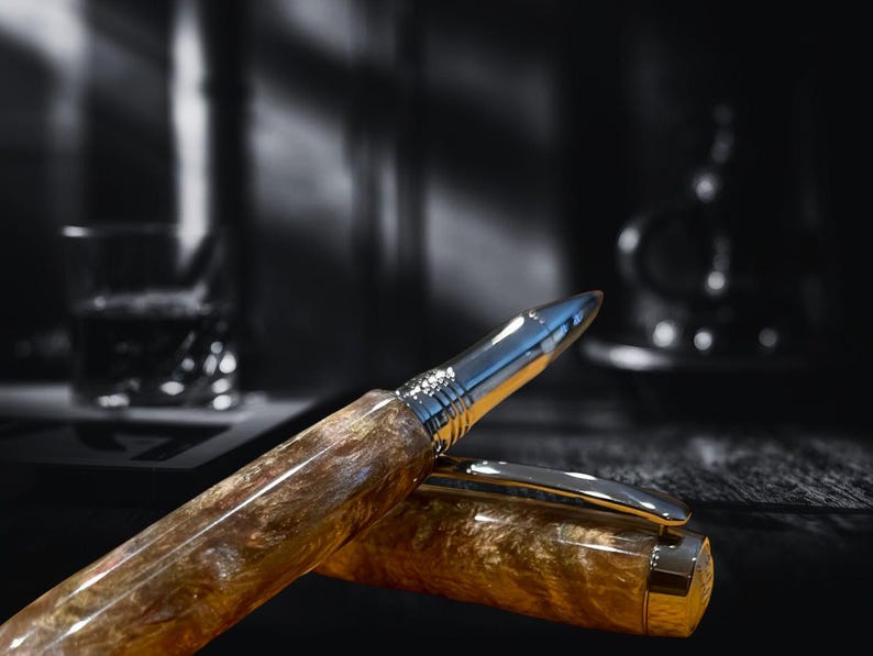 May include: A luxury pen with a silver tip and gold-toned barrel. The pen's barrel has a marbled appearance with brown and gold hues. The pen is partially uncapped, revealing the silver clip and the pen's nib. The background is blurred, with a glass and a dark bottle.