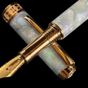 May include: A luxury fountain pen with a gold nib and accents. The pen has a pearlescent white barrel and cap, and a gold clip. The nib is intricately detailed. The pen is set against a black background.