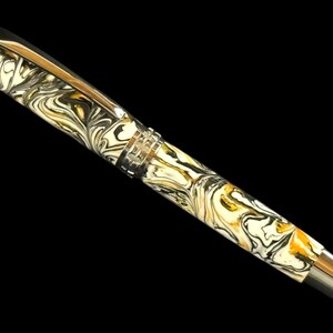 Puede incluir: Una pluma estilogr&aacute;fica de lujo con un dise&ntilde;o veteado en crema, negro y dorado. La pluma presenta un clip y detalles plateados, y un plum&iacute;n plateado. La pluma se encuentra sobre un fondo negro.