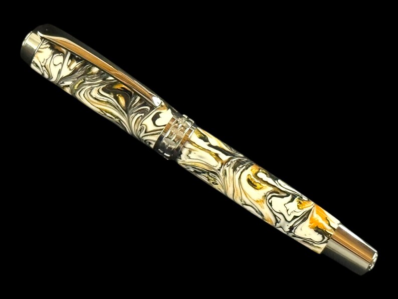 Puede incluir: Una pluma estilogr&aacute;fica de lujo con un dise&ntilde;o veteado en crema, negro y dorado. La pluma presenta un clip y detalles plateados, y un plum&iacute;n plateado. La pluma se encuentra sobre un fondo negro.